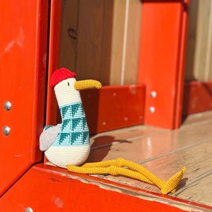 May include: A handmade crochet seagull toy with a red cap, yellow beak, and long yellow legs. The seagull has a white body with a blue and teal patterned design on its chest and gray wings. The toy is sitting on a wooden surface.