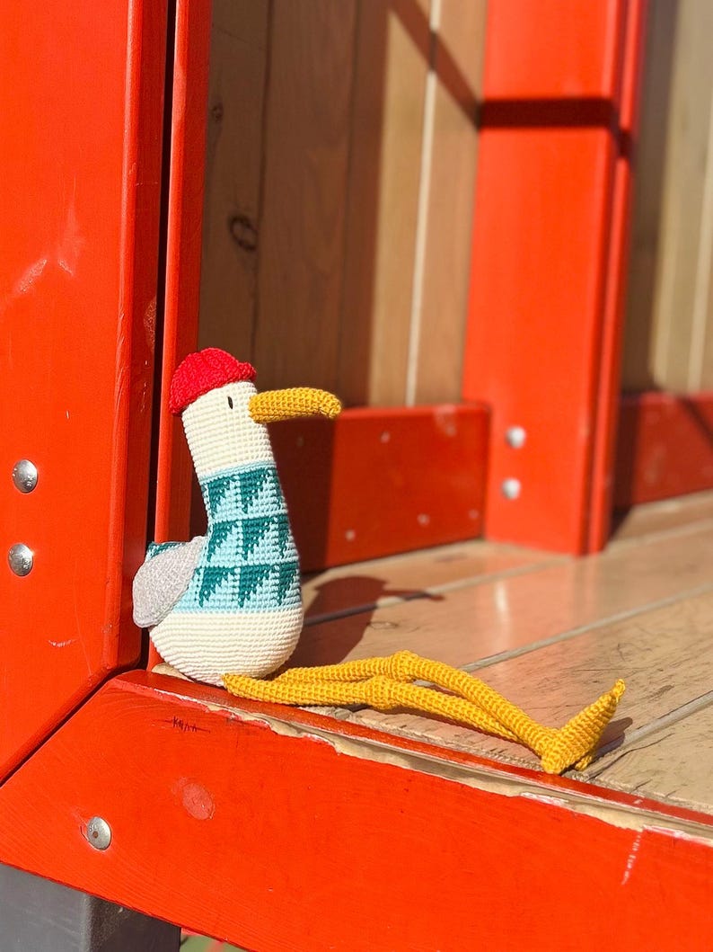 May include: A handmade crochet seagull toy with a red cap, yellow beak, and long yellow legs. The seagull has a white body with a blue and teal patterned design on its chest and gray wings. The toy is sitting on a wooden surface.