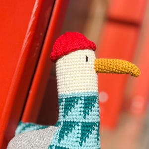 May include: A handmade crochet seagull toy with a red cap, cream-colored head, and a yellow beak. The seagull's neck is adorned with a teal and light blue patterned design. The toy is set against a blurred red background.