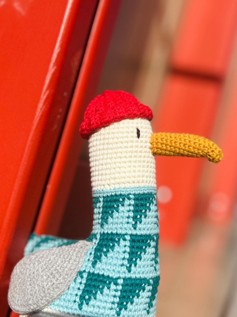 May include: A handmade crochet seagull toy with a red cap, cream-colored head, and a yellow beak. The seagull's neck is adorned with a teal and light blue patterned design. The toy is set against a blurred red background.