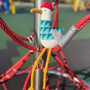 May include: A handmade crochet seagull toy with a red hat, teal and white patterned body, gray wings, and yellow legs. The seagull is perched on a red rope structure, with a blurred playground in the background.