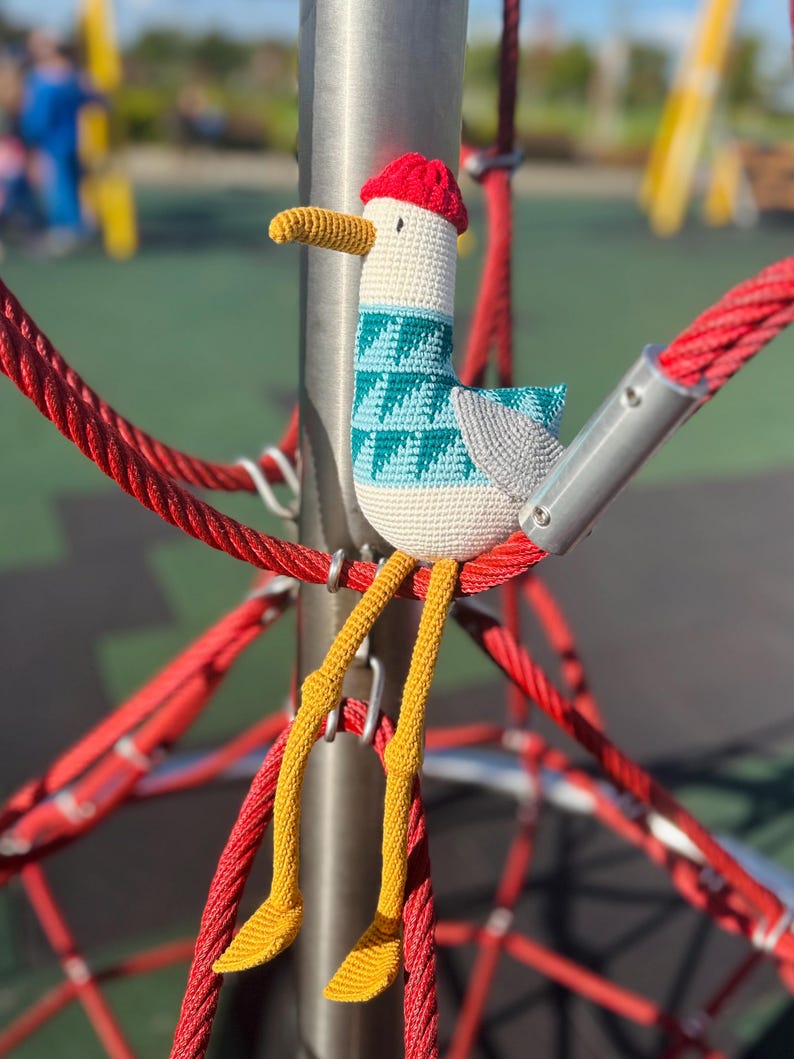 May include: A handmade crochet seagull toy with a red hat, teal and white patterned body, gray wings, and yellow legs. The seagull is perched on a red rope structure, with a blurred playground in the background.