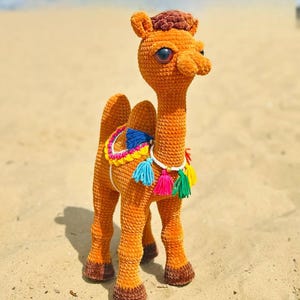 May include: A handmade, orange crochet camel with brown detailing. The camel features large, blue eyes and a colourful saddle with tassels. It is positioned on sand, highlighting the intricate crochet work.