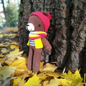 Könnte beinhalten: Ein brauner gehäkelter Teddybär, der einen roten Hut, eine gelbe Weste und einen weiß-rot gestreiften Schal trägt. Der Bär steht auf einem Bett aus gelben Herbstblättern.