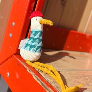 May include: A handmade crochet seagull doll with a red cap, yellow beak and legs, and a teal and white patterned sweater. The seagull is sitting on a wooden surface next to a bright orange structure.