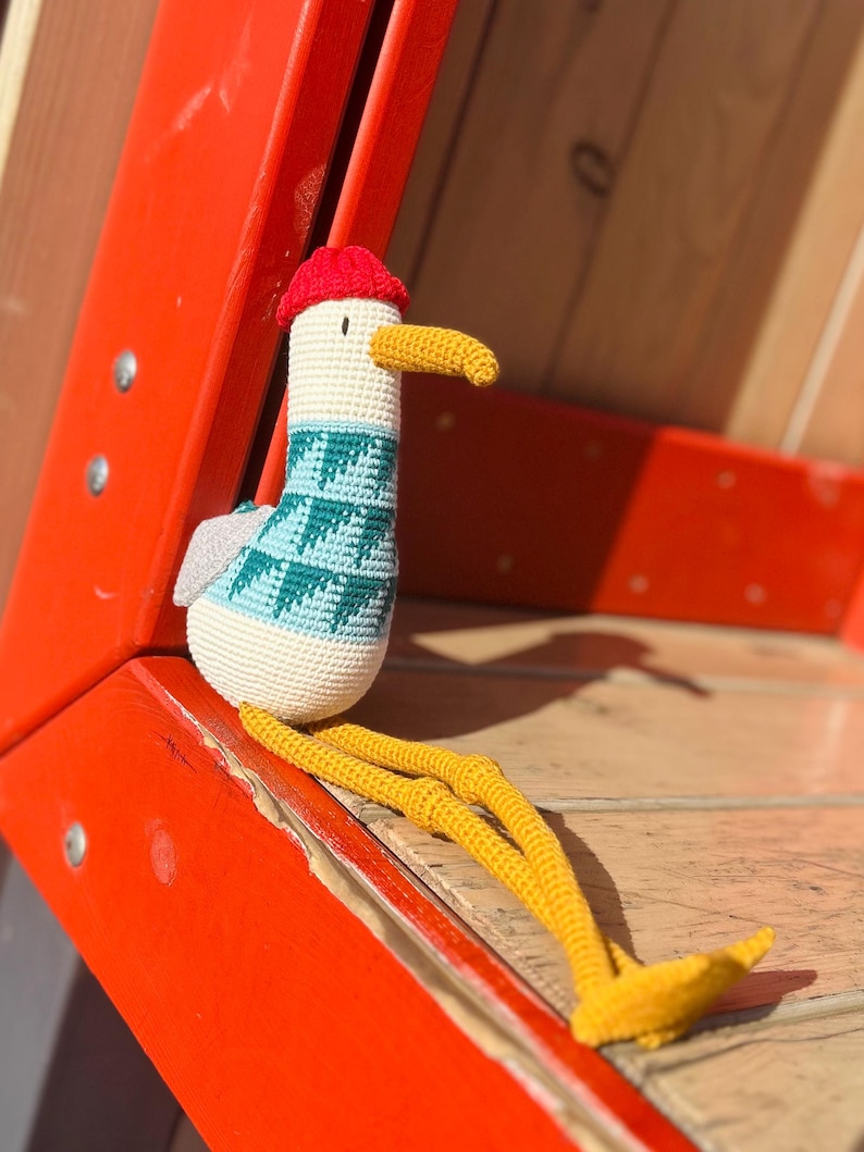 May include: A handmade crochet seagull doll with a red cap, yellow beak and legs, and a teal and white patterned sweater. The seagull is sitting on a wooden surface next to a bright orange structure.
