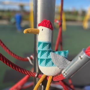 May include: A handmade crochet seagull toy with a red hat, teal and white patterned sweater, and yellow legs. The seagull is perched on a red rope structure, with a blurred playground background.