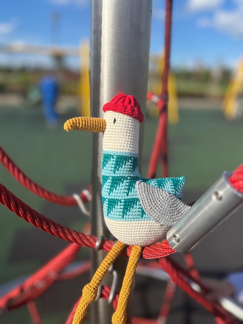 May include: A handmade crochet seagull toy with a red hat, teal and white patterned sweater, and yellow legs. The seagull is perched on a red rope structure, with a blurred playground background.