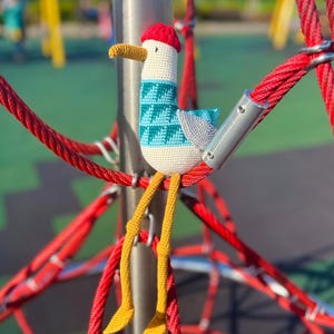 May include: A handmade crochet seagull toy with a red hat, teal and white sweater, and yellow legs. The seagull is perched on a red rope structure, with a blurred playground in the background.