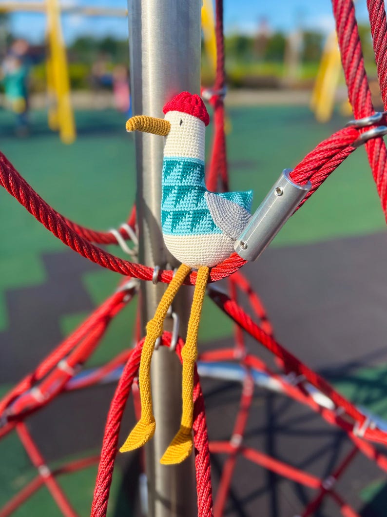 May include: A handmade crochet seagull toy with a red hat, teal and white sweater, and yellow legs. The seagull is perched on a red rope structure, with a blurred playground in the background.