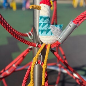 May include: A handmade crochet seagull toy with a red cap, teal and white patterned chest, and yellow legs. The seagull is perched on a red rope structure, showcasing its whimsical design and vibrant colors. The toy is a unique, handcrafted item.