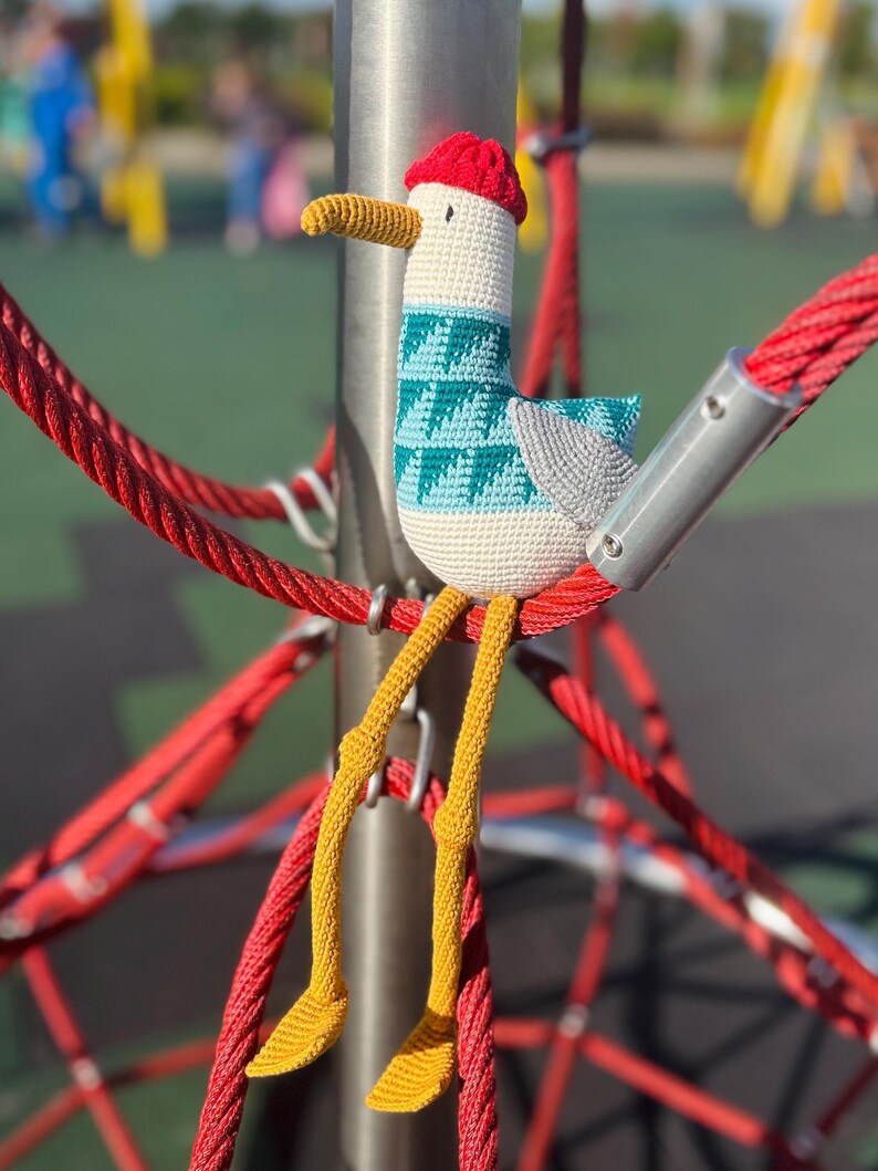 May include: A handmade crochet seagull toy with a red cap, teal and white patterned chest, and yellow legs. The seagull is perched on a red rope structure, showcasing its whimsical design and vibrant colors. The toy is a unique, handcrafted item.