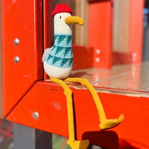 May include: A handmade crochet seagull doll with a red hat, yellow beak and legs, and a teal and white sweater. The seagull is sitting on a red surface.