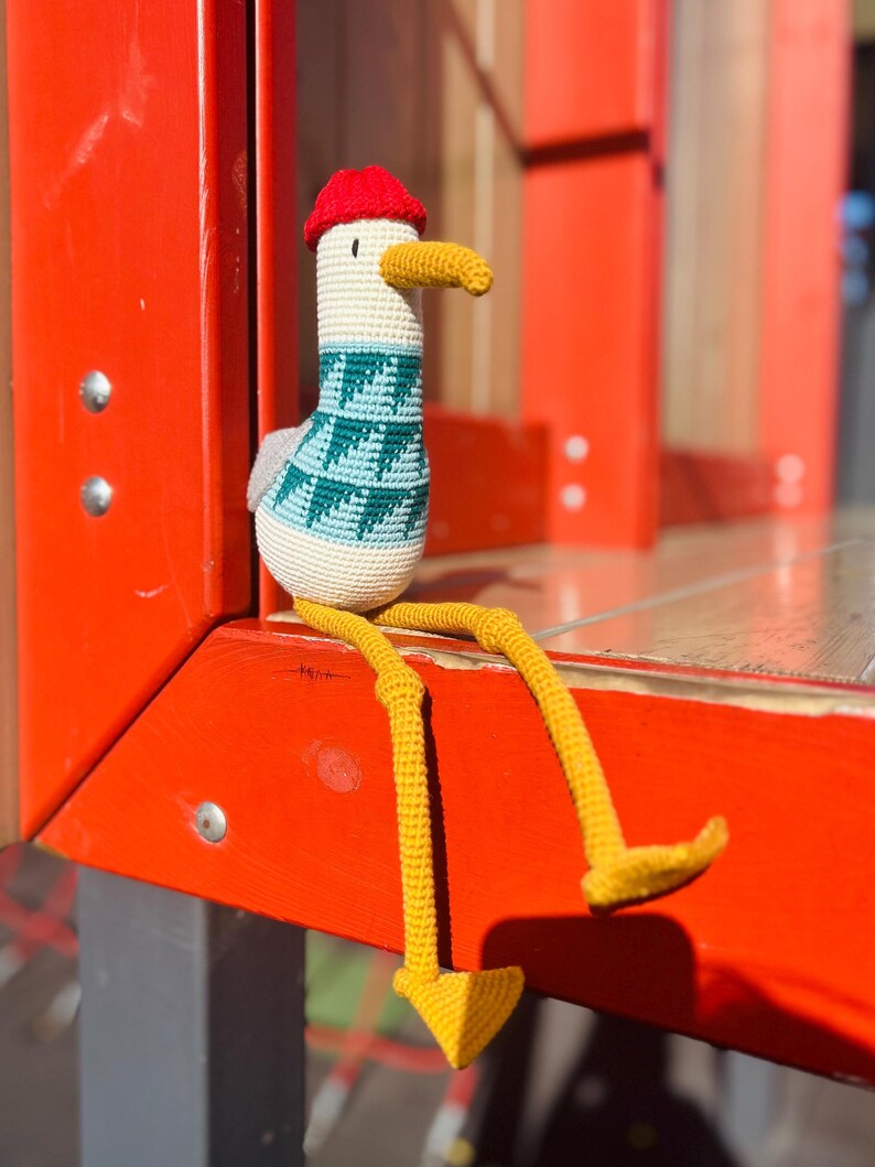 May include: A handmade crochet seagull doll with a red hat, yellow beak and legs, and a teal and white sweater. The seagull is sitting on a red surface.