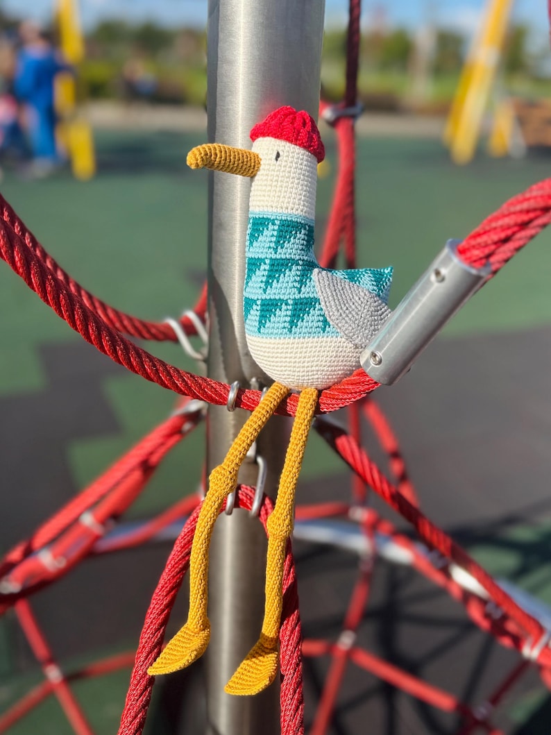 May include: A handmade crochet seagull toy with a red hat, teal and white patterned body, gray wings, and yellow legs. The seagull is perched on a red rope structure, with a blurred playground in the background.