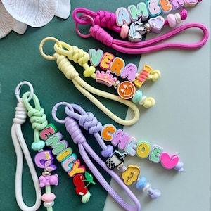 May include: Four colourful wristlet keychains made of braided cord, each featuring custom letter beads spelling names like AMBER, VERA, EMILY, and CHLOE. The keychains are decorated with various charms including a unicorn, crown, smiley face, boba tea, cherry, and heart shapes in pastel and bright tones.