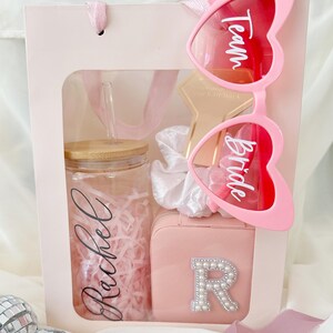 May include: A pink gift bag with a clear window, containing a personalized glass tumbler, a small pink case with a pearl-studded letter "R", a star-shaped accessory, a white scrunchie, and heart-shaped sunglasses with "Team Bride" text.