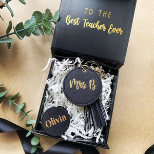 May include: A black gift box with gold lettering that reads "To the Best Teacher Ever". Inside the box is a black keychain with a gold tassel and a black circle with gold lettering that reads "Mrs. B". There is also a smaller black circle with gold lettering that reads "Olivia".