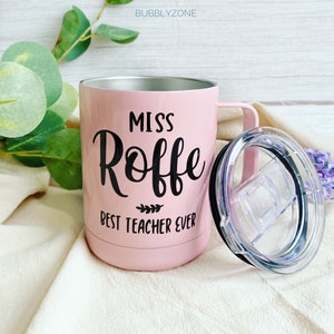 May include: Pink stainless steel travel mug with a black lid and handle. The mug has black text that reads "Miss Roffe Best Teacher Ever".