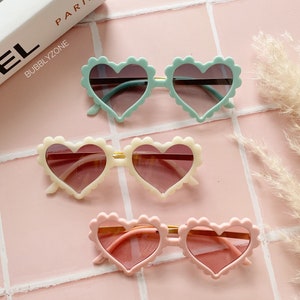 May include: Three pairs of heart-shaped sunglasses with scalloped edges. The sunglasses are in shades of pink, cream, and light blue.