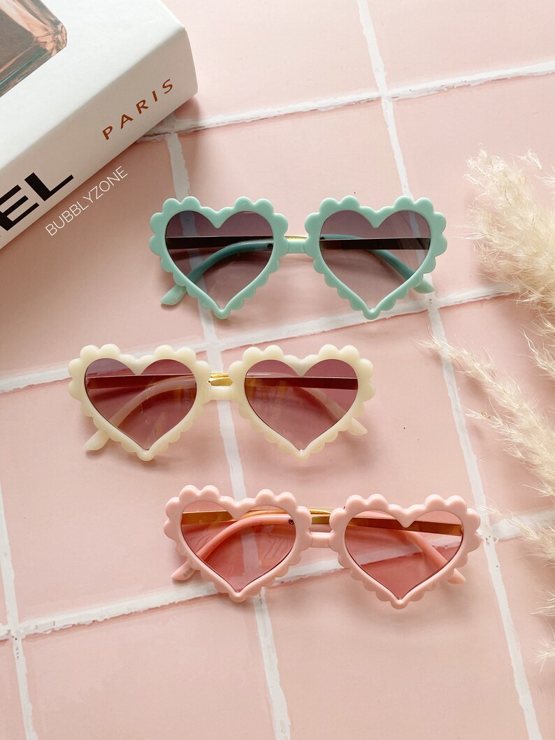May include: Three pairs of heart-shaped sunglasses with scalloped edges. The sunglasses are in shades of pink, cream, and light blue.