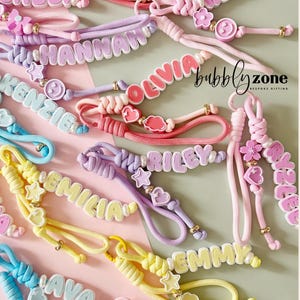 May include: A collection of colourful wrist lanyards featuring personalised names in block letters, accented with small charms like stars, hearts, flowers, and smiley faces. The lanyards are made of braided cord in shades of pink, blue, yellow, and purple.