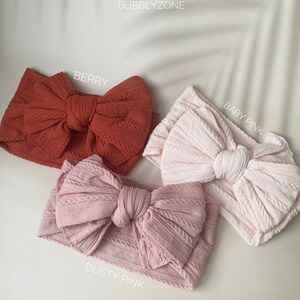 May include: Three fabric headbands with large bows in different colours: berry, baby pink, and dusty pink. The headbands are made of a textured fabric and have a knotted bow in the centre.