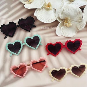 May include: Five pairs of heart-shaped sunglasses in different colours. The sunglasses are all made of plastic and have a scalloped edge. The colours are black, light blue, red, pink, and cream.