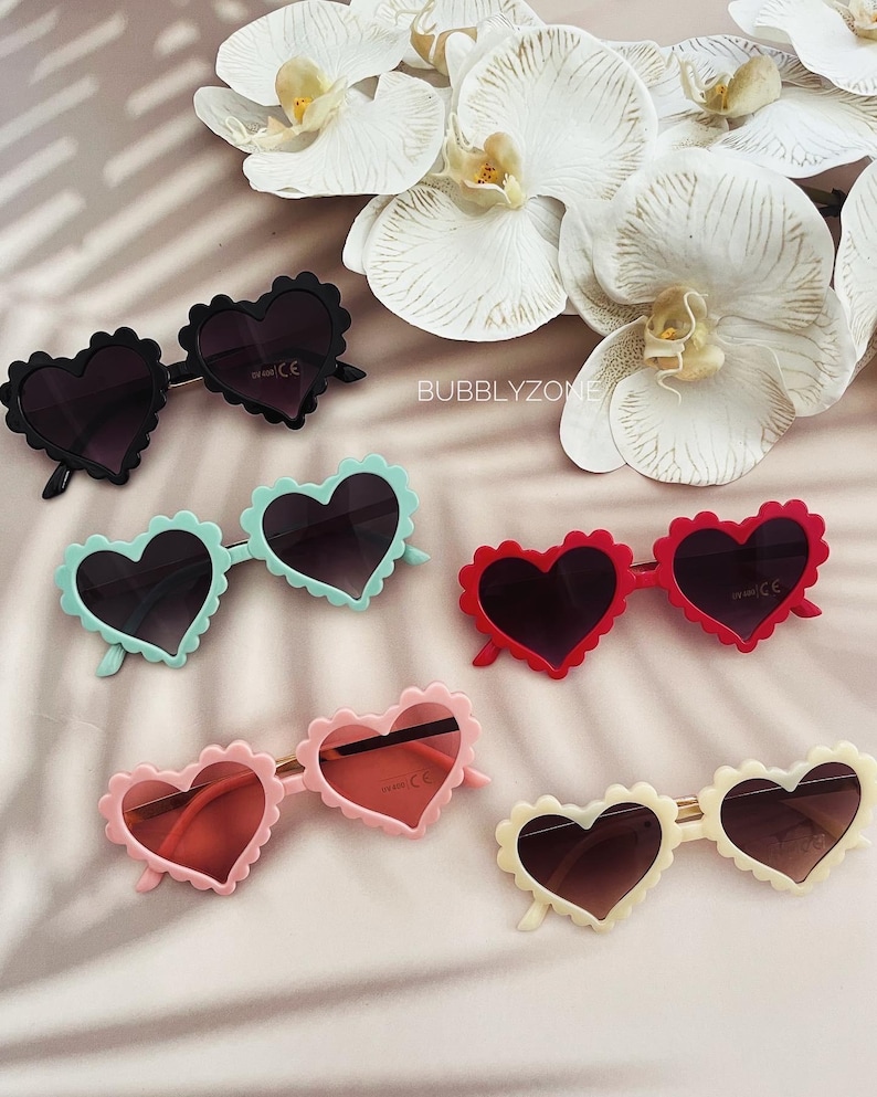 May include: Five pairs of heart-shaped sunglasses in different colours. The sunglasses are all made of plastic and have a scalloped edge. The colours are black, light blue, red, pink, and cream.