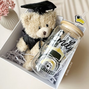May include: A graduation gift set featuring a teddy bear wearing a black graduation cap and a clear glass water bottle with a black and gold graduation design. The bottle has a bamboo lid and a straw with a black and yellow "Graduate" sticker. The text "Emily" is written on the bottle.