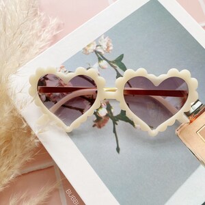 May include: White heart-shaped sunglasses with a scalloped edge and brown lenses. The sunglasses are on a white surface with a floral pattern and the text "BUBBLY_ZONE".