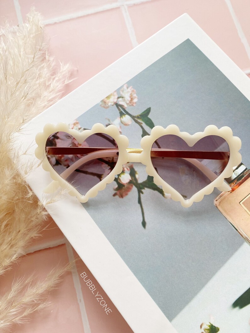 May include: White heart-shaped sunglasses with a scalloped edge and brown lenses. The sunglasses are on a white surface with a floral pattern and the text "BUBBLY_ZONE".