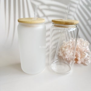 K&ouml;nnte beinhalten: Zwei Glasbecher mit mattem und klarem Finish. Beide haben Holzdeckel und klare Strohhalme. Die Becher stehen auf einer wei&szlig;en Oberfl&auml;che.
