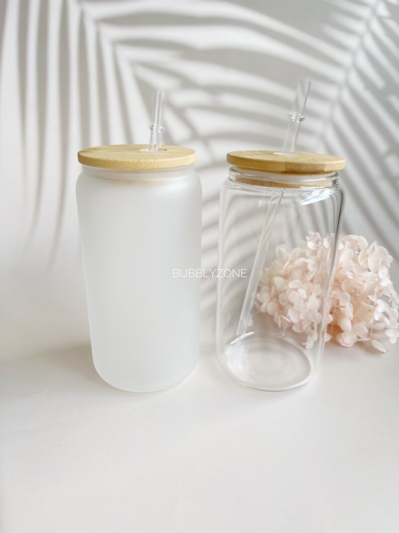 K&ouml;nnte beinhalten: Zwei Glasbecher mit mattem und klarem Finish. Beide haben Holzdeckel und klare Strohhalme. Die Becher stehen auf einer wei&szlig;en Oberfl&auml;che.