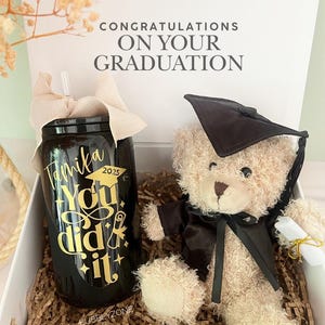 May include: A graduation gift box featuring a black tumbler with gold lettering and a teddy bear wearing a graduation cap. The tumbler reads "Tamila 2025 You did it!" and the box says "Congratulations on Your Graduation."