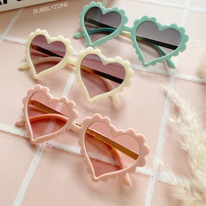 May include: Three pairs of heart-shaped sunglasses with different coloured frames: light blue, cream, and pink. The sunglasses have a scalloped edge and gold-coloured frames.