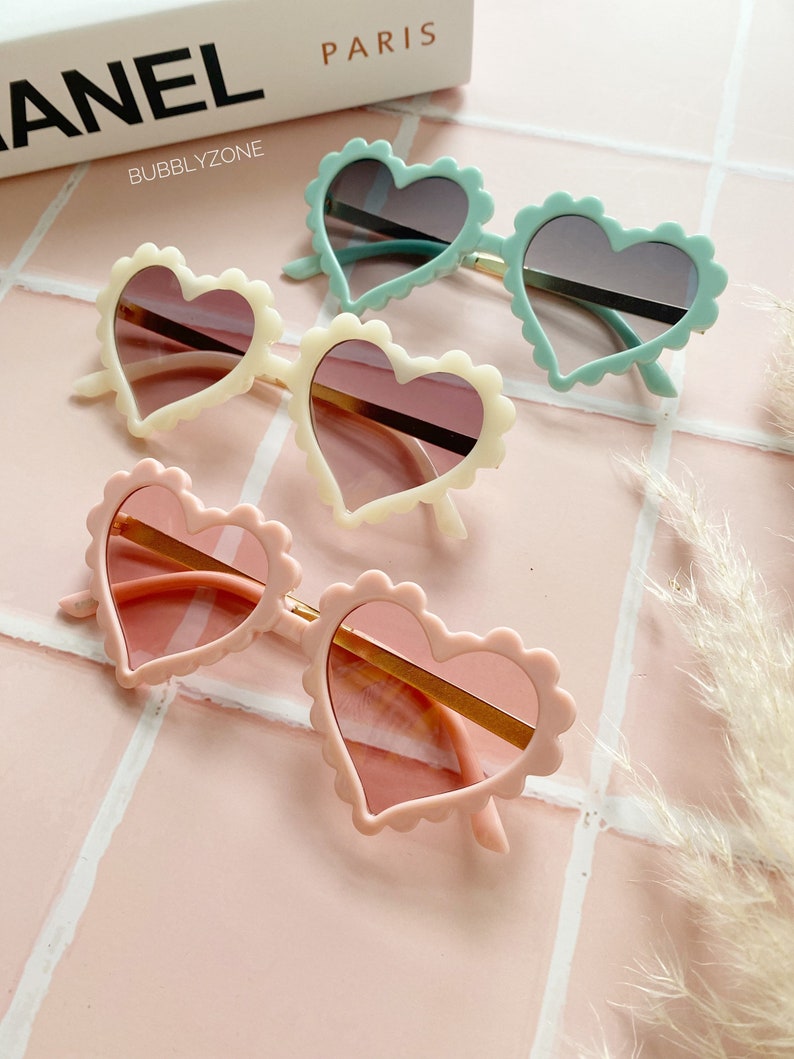 May include: Three pairs of heart-shaped sunglasses with different coloured frames: light blue, cream, and pink. The sunglasses have a scalloped edge and gold-coloured frames.