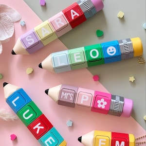 Op de afbeelding: Vijf potloodvormige sleutelhangers in diverse kleuren, elk voorzien van aanpasbare letterblokken. De potloden tonen namen zoals SARA, LEO, LUKE, MRS. P en EM, samen met iconen zoals een smiley en een bloem, afgewerkt met een metalen huls.