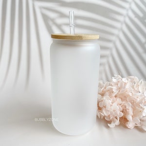 K&ouml;nnte beinhalten: Ein mattierter Glasbecher mit Bambusdeckel und einem klaren Glasstrohhalm. Der Becher steht auf einer wei&szlig;en Oberfl&auml;che mit einem Strau&szlig; getrockneter Blumen.