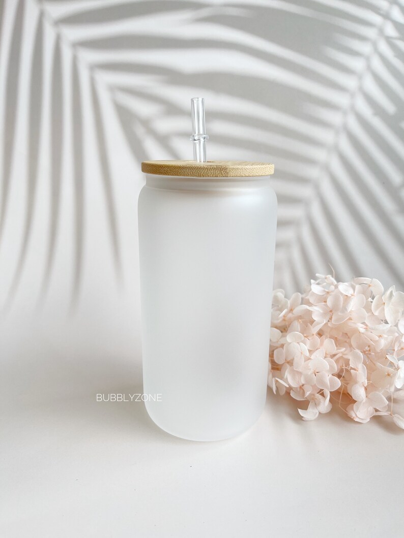 K&ouml;nnte beinhalten: Ein mattierter Glasbecher mit Bambusdeckel und einem klaren Glasstrohhalm. Der Becher steht auf einer wei&szlig;en Oberfl&auml;che mit einem Strau&szlig; getrockneter Blumen.
