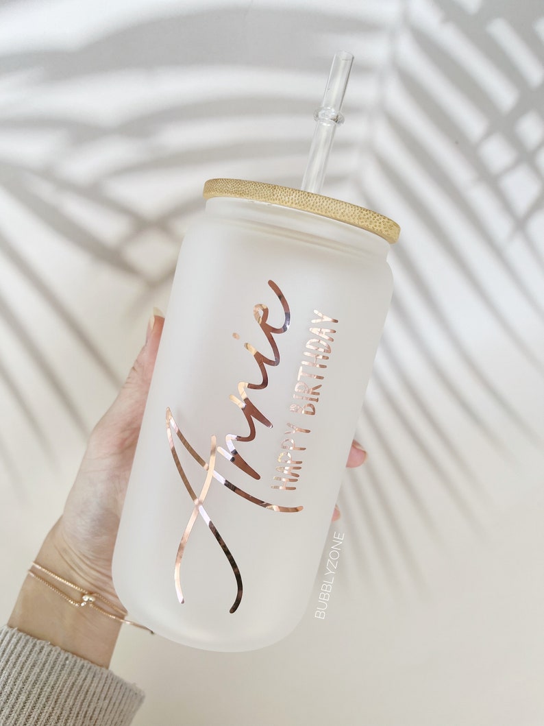 K&ouml;nnte beinhalten: Ein mattierter Glasbecher mit Bambusdeckel und einem klaren Strohhalm. Der Becher hat eine ros&eacute;farbene Aufschrift mit dem Text "Annie Happy Birthday" und den Text "BUBBLYZONE" am Boden.