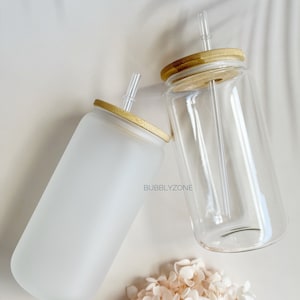 K&ouml;nnte beinhalten: Zwei gl&auml;serne Trinkbecher mit Bambusdeckeln und klaren Trinkhalmen. Ein Becher ist aus mattiertem Glas und der andere aus klarem Glas. Die Becher stehen auf einer wei&szlig;en Oberfl&auml;che mit einem kleinen Strau&szlig; wei&szlig;er Blumen.
