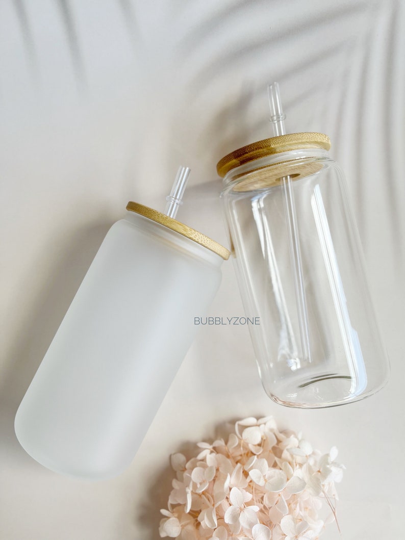 K&ouml;nnte beinhalten: Zwei gl&auml;serne Trinkbecher mit Bambusdeckeln und klaren Trinkhalmen. Ein Becher ist aus mattiertem Glas und der andere aus klarem Glas. Die Becher stehen auf einer wei&szlig;en Oberfl&auml;che mit einem kleinen Strau&szlig; wei&szlig;er Blumen.