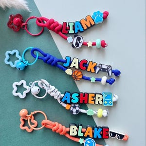 May include: Four personalised keychains featuring colourful paracord loops, star-shaped clips, and small bells. Each keychain displays a name in block letters: LIAM with a panda, JACK with a basketball and controller, ASHER with a soccer ball and handheld game, and BLAKE with a smiley face and sunglasses.