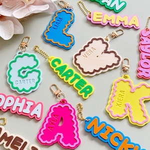 May include: A collection of colourful acrylic keychains featuring various names and letters in diverse fonts and styles. Each keychain includes a metal clasp or chain attachment. The names displayed include Brian, Emma, Liam, Vera, Carter, Kiera, Sophia, Ayla, Nicky, and Amelia.