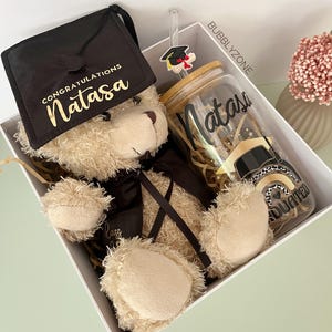 May include: A white gift box containing a brown teddy bear, a black graduation cap, and a clear glass with a bamboo lid and straw. The glass has the name "Natasa" written on it and a graduation cap sticker. The box is filled with brown shredded paper. The text on the graduation cap reads "Congratulations Natasa".