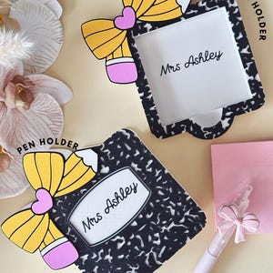 May include: A black and white composition notebook patterned sticky note holder and a matching pen holder, both featuring a yellow pencil bow design with a pink heart center. Each item is personalized with the text Mrs Ashley in black script.