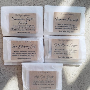 May include: Five small white paper bags with brown labels. Each bag contains a 1 ounce mini soap bar. The labels have the text "The Sugar Apothecary" and a different scent name: "Cinnamon Sugar Donuts", "Sugared Lemonade", "Lemon Blueberry Cake", "Cold Brew Coffee", and "Apple Cider Donuts".