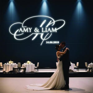 以下が含まれることがあります： 黒い壁の前に、花嫁と新郎がキスをしています。壁には「AMY & LIAM」と「14.06.2018」と書かれた白いネオンサインがあります。