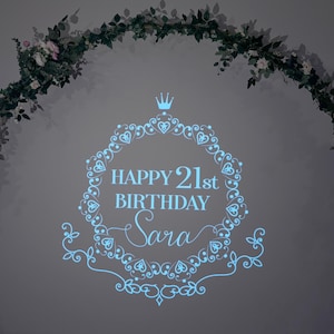 May include: A blue and white projected image of a decorative frame with the text "Happy 21st Birthday Sara" in a light blue font. The image is projected onto a white wall with a green garland of leaves and flowers around the top.